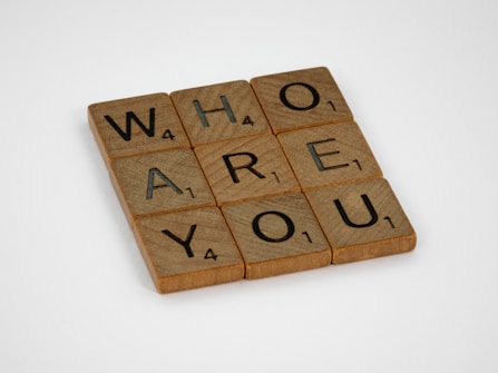 Wooden letter tiles are placed on a white surface forming the words 'WHO ARE YOU'. Each tile has a letter along with a number in the bottom right corner, similar to a Scrabble game.