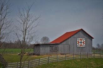 A rustic barn with a grey wooden exterior and a vibrant red roof stands amidst a rural landscape. The barn features a large, colorful quilt-like pattern on its side. Surrounding the barn is a wooden fence, bare trees, and a grassy field, indicating a late autumn or early spring setting.