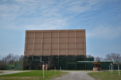 A large, old drive-in movie theater screen stands prominently in an open area, surrounded by sparse trees. The screen is supported by a wooden structure with faded paint, indicating age and disuse. There's a small building nearby and several lamp posts along a narrow road leading up to the screen.