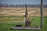 An old, rustic water pump stands on a concrete slab in the foreground of a rural setting. Surrounding the pump is a field of dry, harvested land extending towards the horizon. A red barn and some trees are visible in the distance underneath a blue sky. The pump features a large wheel and metal components giving it an antique appearance.