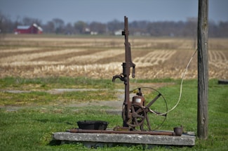 An old, rustic water pump stands on a concrete slab in the foreground of a rural setting. Surrounding the pump is a field of dry, harvested land extending towards the horizon. A red barn and some trees are visible in the distance underneath a blue sky. The pump features a large wheel and metal components giving it an antique appearance.