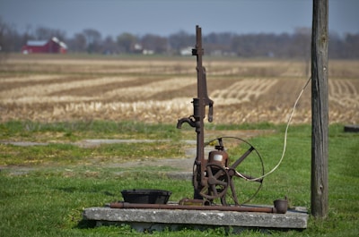 An old, rustic water pump stands on a concrete slab in the foreground of a rural setting. Surrounding the pump is a field of dry, harvested land extending towards the horizon. A red barn and some trees are visible in the distance underneath a blue sky. The pump features a large wheel and metal components giving it an antique appearance.