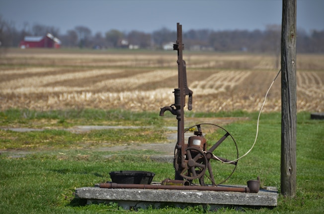 An old, rustic water pump stands on a concrete slab in the foreground of a rural setting. Surrounding the pump is a field of dry, harvested land extending towards the horizon. A red barn and some trees are visible in the distance underneath a blue sky. The pump features a large wheel and metal components giving it an antique appearance.
