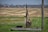 An old, rustic water pump stands on a concrete slab in the foreground of a rural setting. Surrounding the pump is a field of dry, harvested land extending towards the horizon. A red barn and some trees are visible in the distance underneath a blue sky. The pump features a large wheel and metal components giving it an antique appearance.