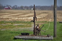 An old, rustic water pump stands on a concrete slab in the foreground of a rural setting. Surrounding the pump is a field of dry, harvested land extending towards the horizon. A red barn and some trees are visible in the distance underneath a blue sky. The pump features a large wheel and metal components giving it an antique appearance.