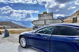 A blue car is parked beside a road sign on a mountainous pass with a stunning view of clouds and distant mountain peaks. The sign indicates various directions and distances, including La Bonneval-Sur-Arc and Lanslebourg, with an altitude marking of 2770 meters. The landscape features rocky terrain and patches of grass with clear skies overhead.
