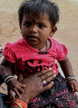 A young child with short, dark hair and a small decorative mark on their forehead is wearing a bright pink top with black lettering and dark jeans. They are held gently by an adult's hand adorned with rings and bracelets, complementing the child’s bracelets. The background is neutral and unobtrusive.