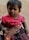 A young child with short, dark hair and a small decorative mark on their forehead is wearing a bright pink top with black lettering and dark jeans. They are held gently by an adult's hand adorned with rings and bracelets, complementing the child’s bracelets. The background is neutral and unobtrusive.