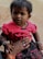 A young child with short, dark hair and a small decorative mark on their forehead is wearing a bright pink top with black lettering and dark jeans. They are held gently by an adult's hand adorned with rings and bracelets, complementing the child’s bracelets. The background is neutral and unobtrusive.