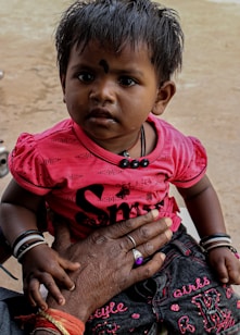 A young child with short, dark hair and a small decorative mark on their forehead is wearing a bright pink top with black lettering and dark jeans. They are held gently by an adult's hand adorned with rings and bracelets, complementing the child’s bracelets. The background is neutral and unobtrusive.