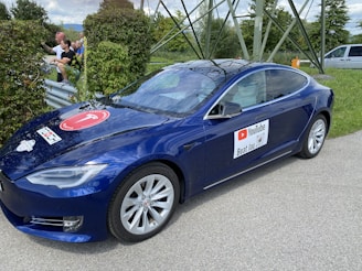 A blue Tesla car is parked near a grassy area with trees and a metal structure in the background. The car has a sign on the door featuring a YouTube logo and text. On the hood, there is another circular logo. The day is partly cloudy, with shadows visible on the car. In the background, a few people are visible near the roadside, partially obscured by bushes.