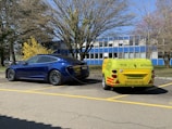 A blue electric car is parked in a lot and is connected to a small yellow charging trailer labeled with eCharge. The setting includes a few trees without leaves, and a building with blue and white panels in the background.