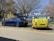 A blue electric car is parked in a lot and is connected to a small yellow charging trailer labeled with eCharge. The setting includes a few trees without leaves, and a building with blue and white panels in the background.