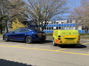 A blue electric car is parked in a lot and is connected to a small yellow charging trailer labeled with eCharge. The setting includes a few trees without leaves, and a building with blue and white panels in the background.