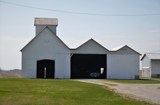 A large, white barn with a tin roof stands prominently on a grassy landscape. The barn has open doors revealing a vehicle inside. Adjacent structures with similar tin roofs are visible in the background. A dirt path leads to the barn, curving gently from the foreground to the background.