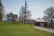 A picturesque rural landscape featuring a white two-story house with a porch, surrounded by trees. To the right is a red barn with a metal roof, set against a backdrop of leafless trees. The foreground shows a lush, well-manicured lawn and a gravel driveway leading to the barn.