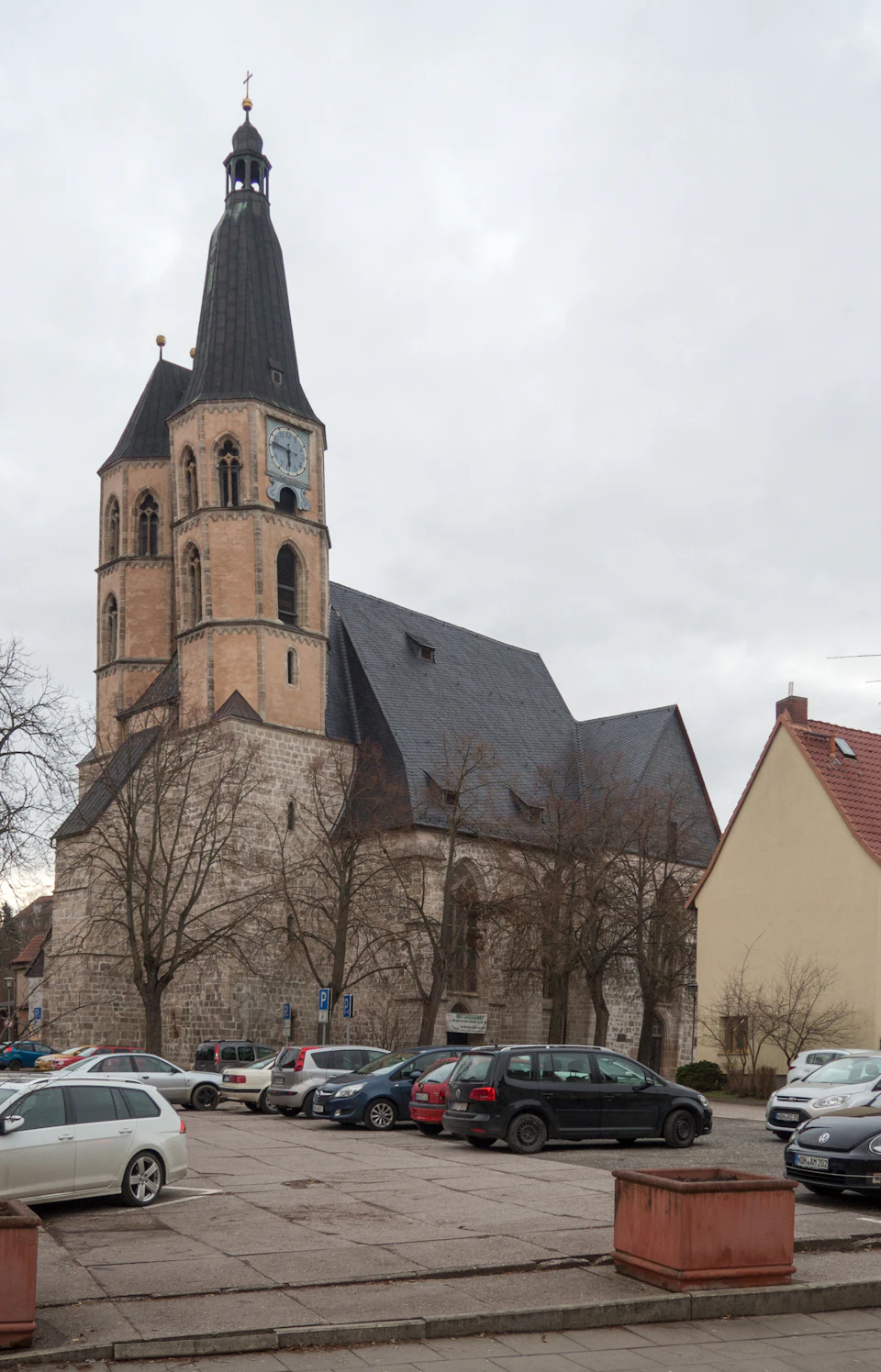 The image depicts a large, Gothic-style church with a tall steeple and a clock face on each side. The church is situated within an urban setting, surrounded by trees that provide some natural greenery to the scene. In front of the church, there are several cars parked along the street, indicating that this location may be easily accessible for visitors or locals alike.