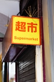 A bright yellow sign with red Chinese characters and the word 'Supermarket' in English below. It is mounted on a metal structure attached to a building facade.