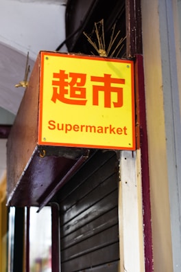 A bright yellow sign with red Chinese characters and the word 'Supermarket' in English below. It is mounted on a metal structure attached to a building facade.