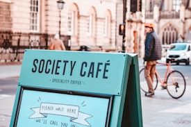 A prominent sign displays the words 'Society Caf&eacute; Speciality Coffee' in a city setting. In the background, a blurred pedestrian dressed in a helmet and jacket walks past a brick building while pushing an orange bicycle. The scene captures an urban, relaxed atmosphere.
