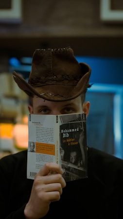 A person wearing a brown cowboy hat holds up a book titled 'Le m&eacute;tier &agrave; tisser' by Mohammed Dib, partially covering their face. The background is softly blurred, suggesting an indoor setting, possibly a library or bookstore.