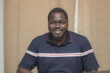A man with a beard is smiling while wearing a navy blue polo shirt with red and white stripes. He is standing indoors in front of a beige background.
