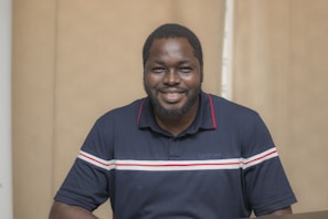 A man with a beard is smiling while wearing a navy blue polo shirt with red and white stripes. He is standing indoors in front of a beige background.
