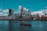 A cityscape featuring a prominent, modern skyline with unique skyscrapers standing beside historic buildings. A river flows in the foreground with small boats on its surface, and fluffy clouds are scattered across the bright blue sky.