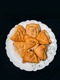 A white plate with an artistic floral design holds several triangular pastries. The pastries have a golden-brown baked appearance with a textured surface and are neatly arranged on the plate.
