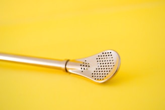 A metallic utensil with a perforated, spatula-like head is placed against a solid yellow background. The utensil has a sleek and shiny appearance, and its design features small holes for straining or aeration purposes.