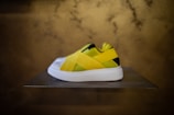 A sneaker with a unique design, featuring vibrant yellow and black straps crossing over a white base. The sneaker is displayed on a flat, raised surface against a blurred, textured gold background.