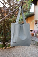 A blue tote bag is hanging from a tree branch in an outdoor setting with a pebbled ground. The background consists of blurred greenery and part of a building or house with visible windows and a potted plant.