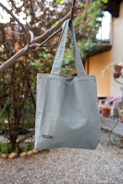 A blue tote bag is hanging from a tree branch in an outdoor setting with a pebbled ground. The background consists of blurred greenery and part of a building or house with visible windows and a potted plant.