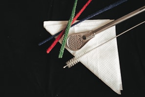 A metal perforated strainer rests on a white paper napkin, accompanied by three colored pipe cleaners in red, green, and blue. A cleaning brush lies parallel to the strainer, all set against a dark background.