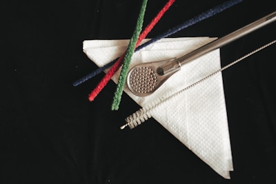 A metal perforated strainer rests on a white paper napkin, accompanied by three colored pipe cleaners in red, green, and blue. A cleaning brush lies parallel to the strainer, all set against a dark background.