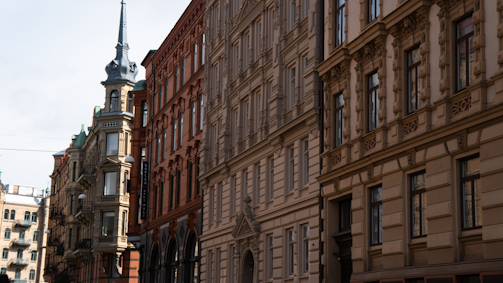A row of historic buildings with ornate architectural details. The facades feature a combination of brick and stucco, with decorative elements around the windows. The street scene includes a pointed tower on one of the buildings and several windows reflecting light.