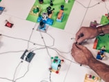 Several hands work on arranging colorful Lego pieces and wires on a white surface. The scene appears to involve multiple platforms made of Lego with miniature models and figures placed on them. The wires are spread across and seem to interconnect the Lego pieces.