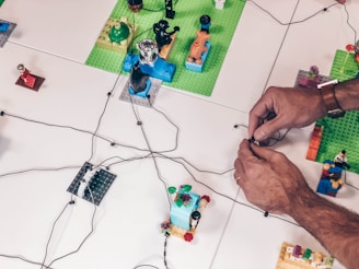 Several hands work on arranging colorful Lego pieces and wires on a white surface. The scene appears to involve multiple platforms made of Lego with miniature models and figures placed on them. The wires are spread across and seem to interconnect the Lego pieces.