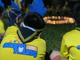 Children dressed in yellow uniforms with blue designs sit on grass, with one wearing a blue paper crown labeled with a name. A circle of soft candlelight glows in the background.