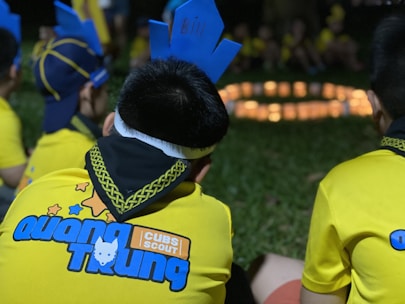 Children dressed in yellow uniforms with blue designs sit on grass, with one wearing a blue paper crown labeled with a name. A circle of soft candlelight glows in the background.