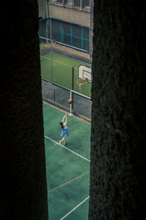 A young person is playing basketball on a green outdoor court, attempting a shot at the hoop. The court is enclosed by a net and surrounded by buildings, with the perspective framed between two large dark pillars.