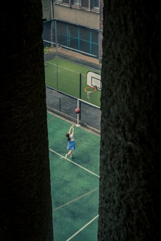 A young person is playing basketball on a green outdoor court, attempting a shot at the hoop. The court is enclosed by a net and surrounded by buildings, with the perspective framed between two large dark pillars.