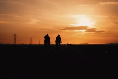 Sunset silhouette of cyclists riding along an open road, with city lights ahead.