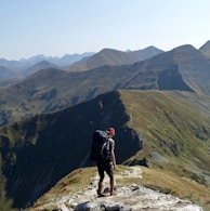 A lone hiker stands on a rocky trail atop a mountain ridge, wearing a large backpack and a red cap. The expansive view showcases a series of rugged, green and brown mountains under a clear blue sky.