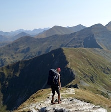 A lone hiker stands on a rocky trail atop a mountain ridge, wearing a large backpack and a red cap. The expansive view showcases a series of rugged, green and brown mountains under a clear blue sky.