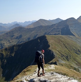 A lone hiker stands on a rocky trail atop a mountain ridge, wearing a large backpack and a red cap. The expansive view showcases a series of rugged, green and brown mountains under a clear blue sky.