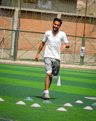 A person in sports clothing is running on a green artificial turf with white and red cones arranged in a pattern. The individual appears to be participating in a sports drill or exercise. The background includes a brick wall and netting consistent with an outdoor sports facility.