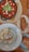 A wooden table displays a rustic meal featuring a bowl of red tomato-based stew topped with sliced avocado, crumbled cheese, and herbs. Beside it, a plate holds slices of toasted bread. Another bowl contains a creamy white dip garnished with herbs and a drizzle of oil.