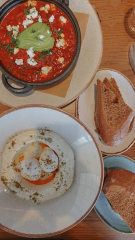 A wooden table displays a rustic meal featuring a bowl of red tomato-based stew topped with sliced avocado, crumbled cheese, and herbs. Beside it, a plate holds slices of toasted bread. Another bowl contains a creamy white dip garnished with herbs and a drizzle of oil.