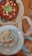 A wooden table displays a rustic meal featuring a bowl of red tomato-based stew topped with sliced avocado, crumbled cheese, and herbs. Beside it, a plate holds slices of toasted bread. Another bowl contains a creamy white dip garnished with herbs and a drizzle of oil.